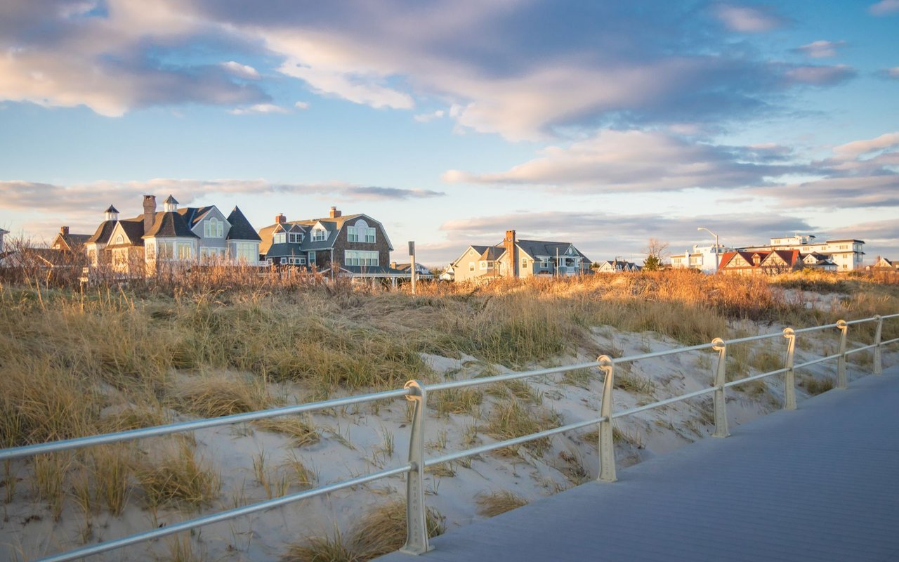 Coastal homes behind grassy dunes, viewed from a boardwalk with a metal railing.