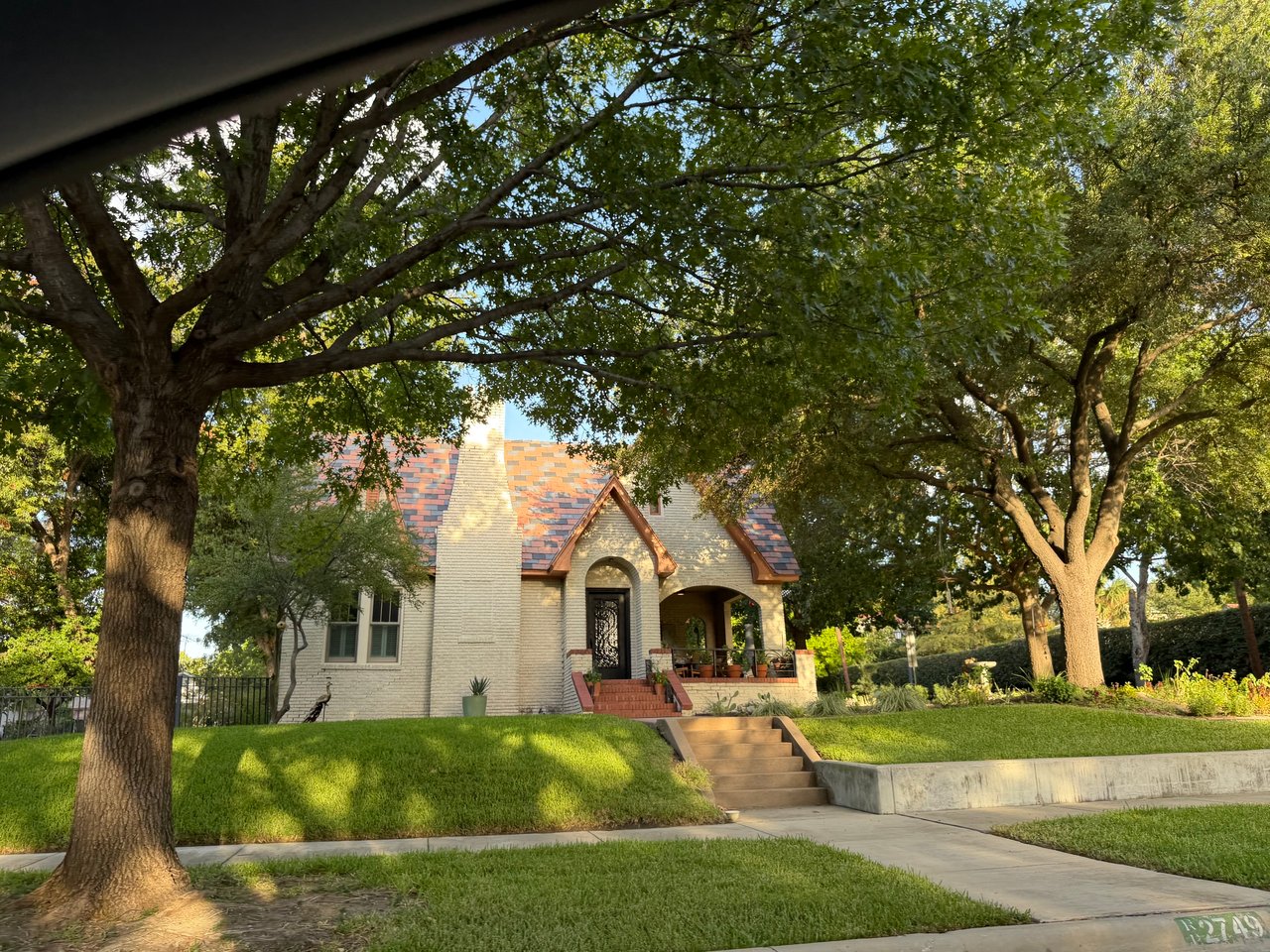 Light brick home with a steep gabled roof and deep porch beneath mature shade trees.