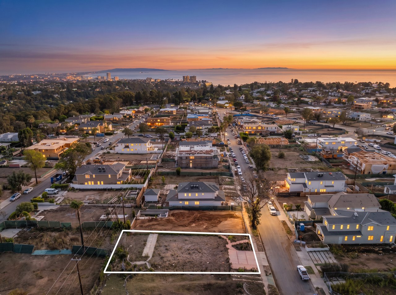Aerial view of a vacant lot outlined in white, with neighborhood and ocean at sunset.