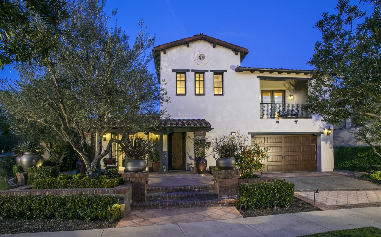 Two-story stucco home with a balcony, tiled roof, and landscaped front entry at dusk.