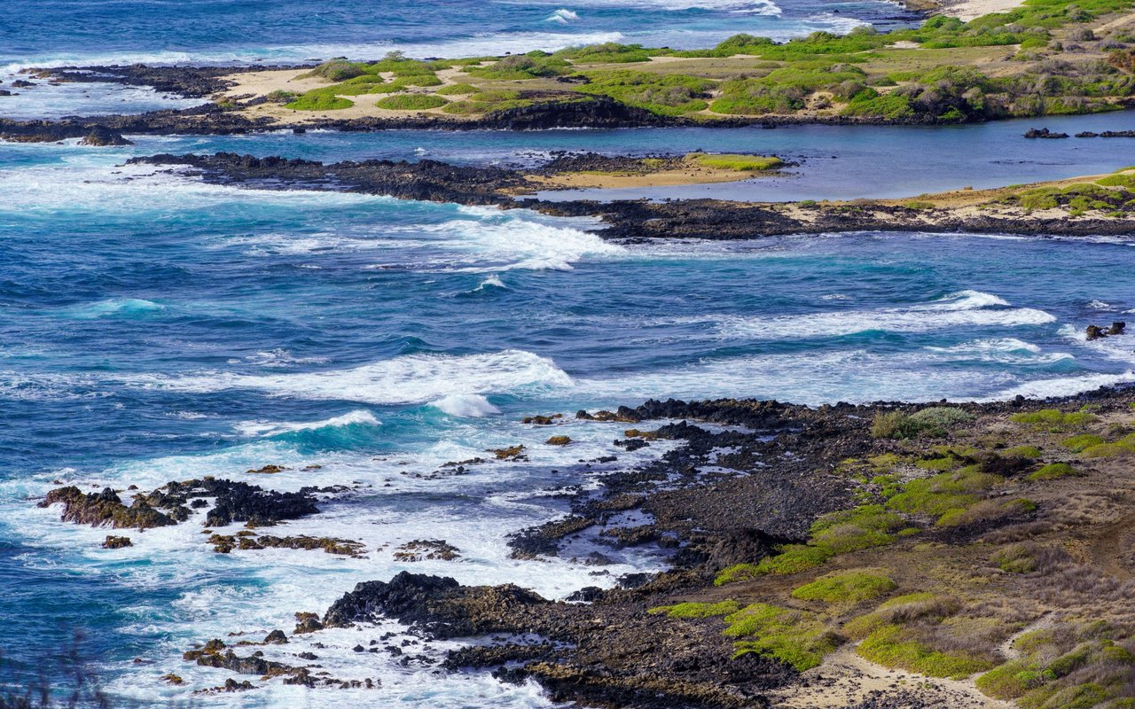 Paiko Lagoon’s calm water sits behind a rocky shore as ocean waves break in the foreground.