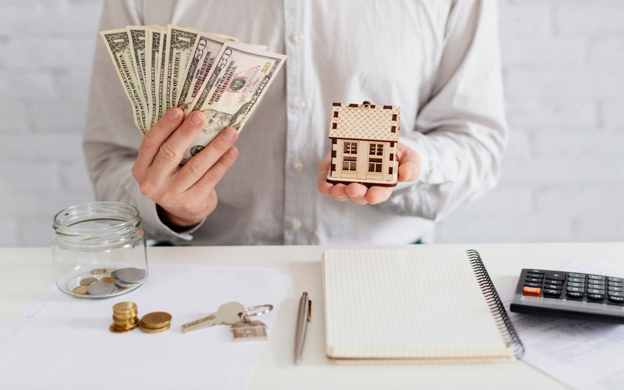 Hands hold cash and a small wooden house model above a desk with keys and a coin jar.