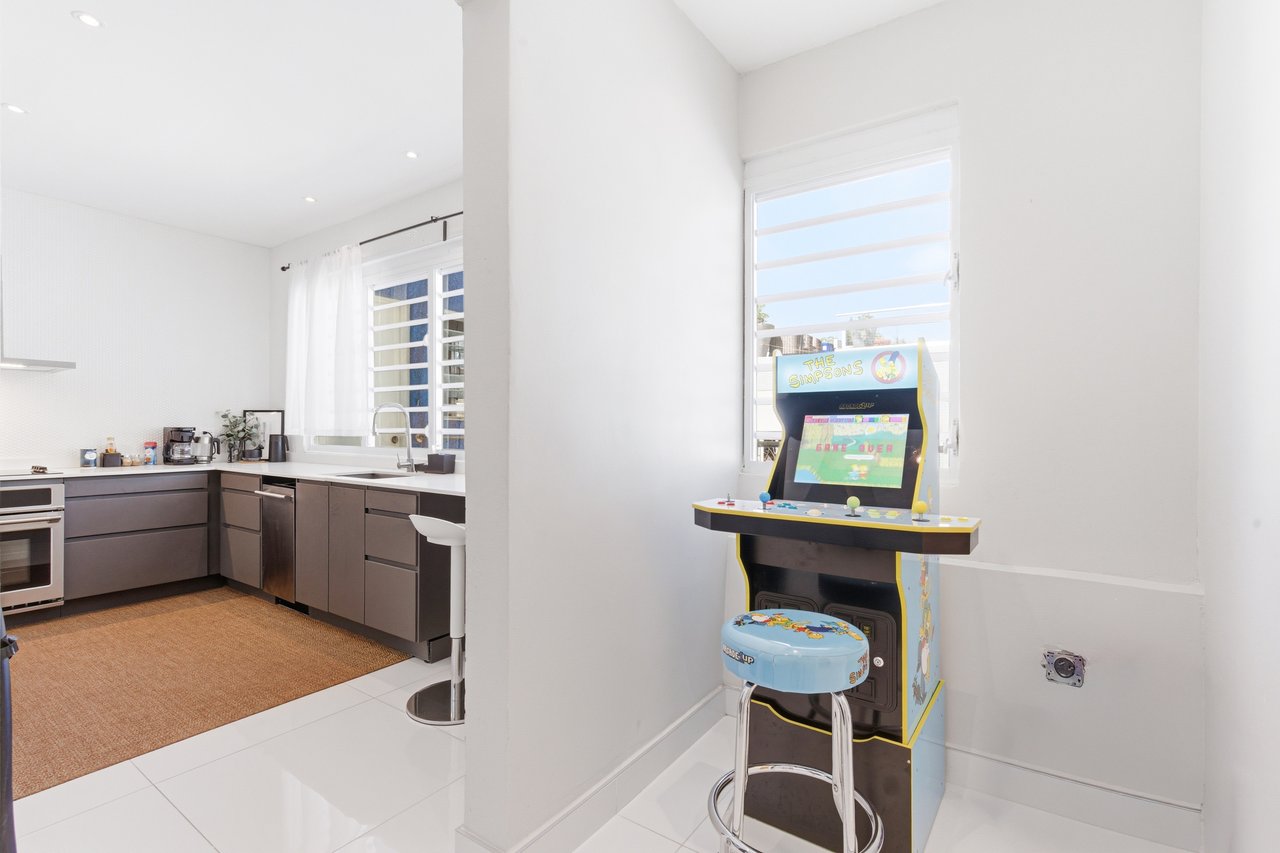 Modern kitchen beside a Simpsons arcade game and stool by a large window.