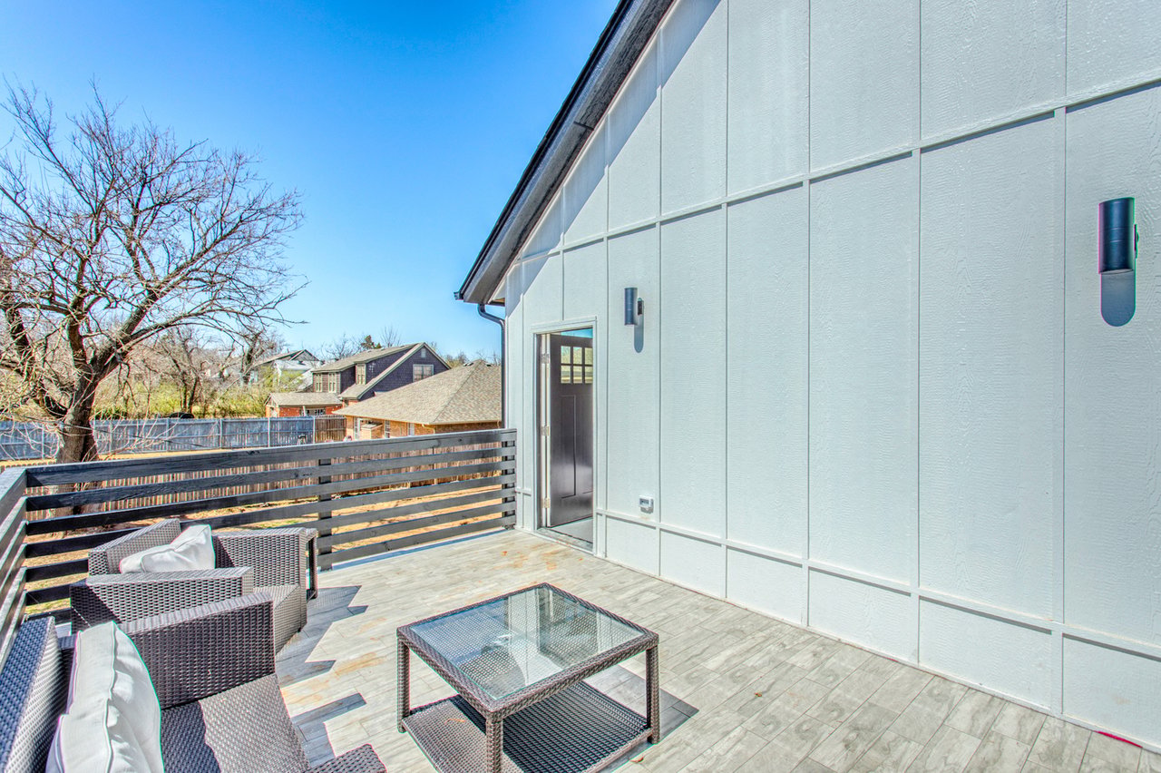 Raised deck with gray outdoor seating and glass-top table beside a light-colored house.