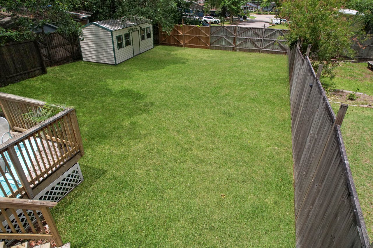 A spacious backyard with a vibrant green lawn, bordered by wooden fences. A wooden deck is on the left, and a storage shed is near the back fence.