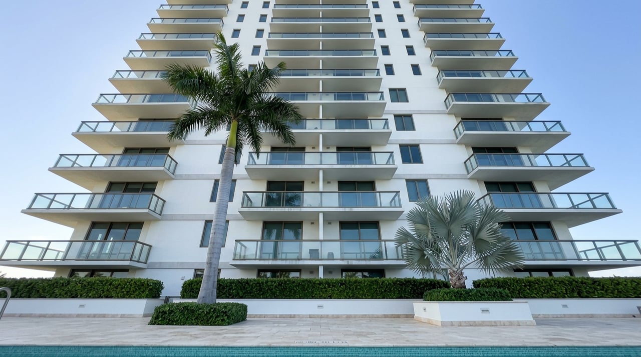 Waterfront condominium tower with stacked glass balconies, framed by royal and silver-blue palms above a pool deck.
