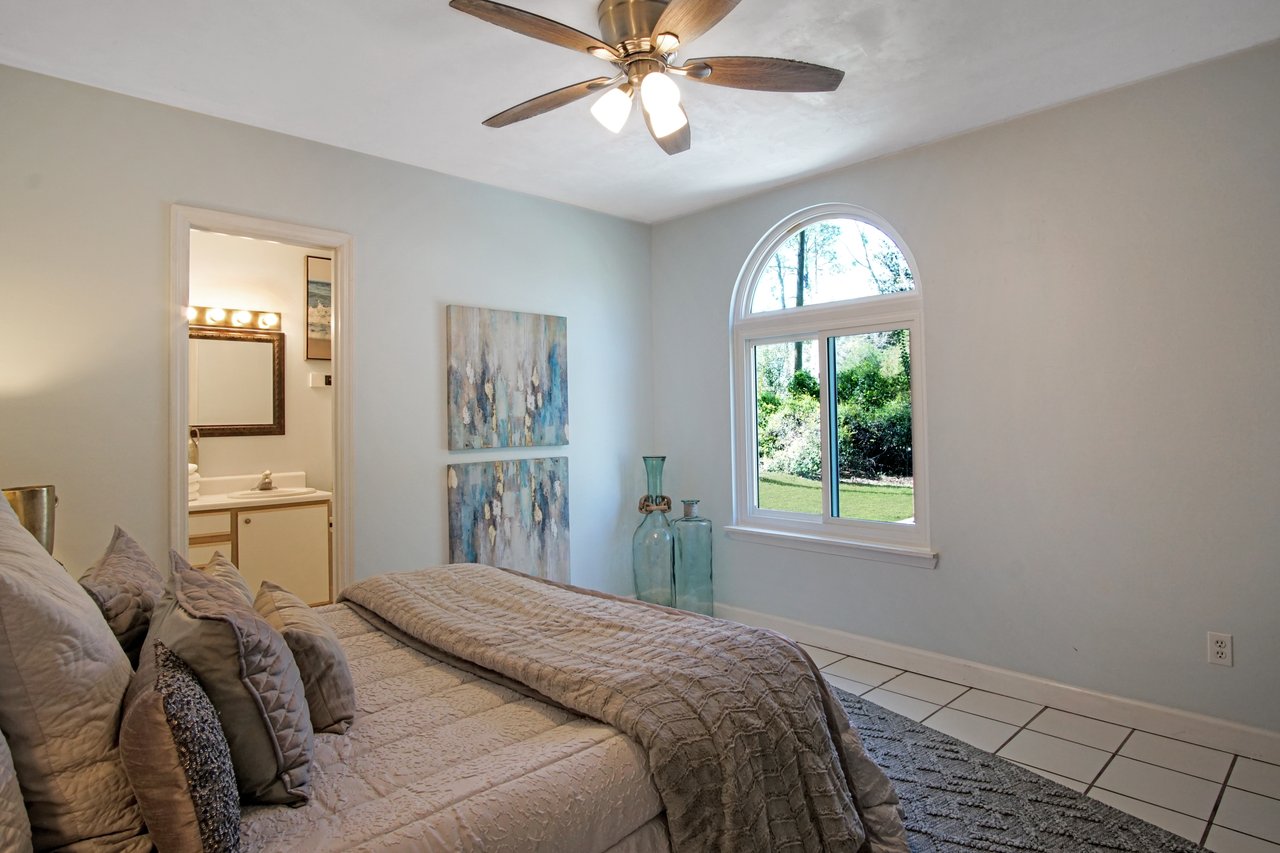 Cozy bedroom with a soft, quilted bedspread and pillows under a ceiling fan. An arched window offers a view of the lush green outdoors.Cozy bedroom with a soft, quilted bedspread and pillows under a ceiling fan. An arched window offers a view of the lush green outdoors.