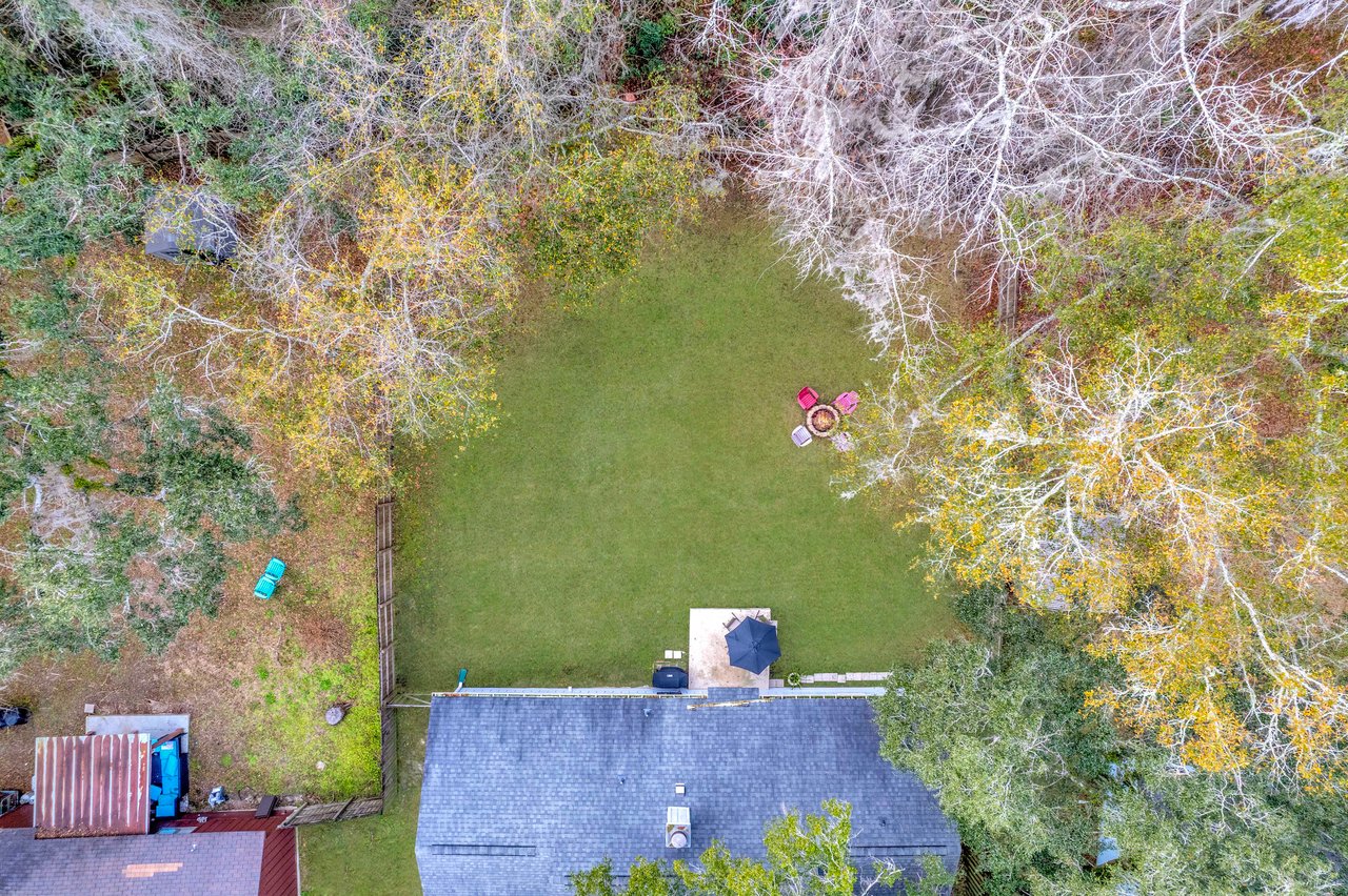 Aerial view of a backyard with lush green grass. A house with a dark roof is at the bottom. Four colorful chairs surround a fire pit in the yard's corner, bordered by trees with autumn leaves.