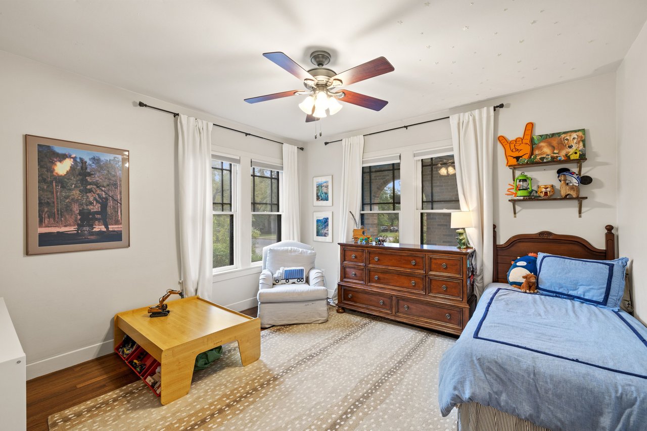 Bedroom with a blue-covered bed, wooden dresser, play table, and bright windows.