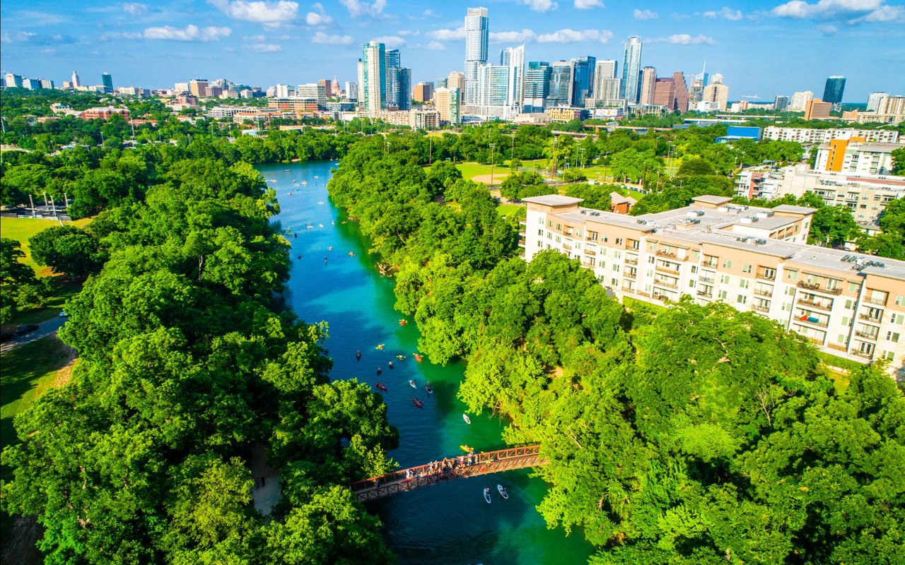 A tree-lined river and park with a pedestrian bridge, apartments, and downtown skyline.