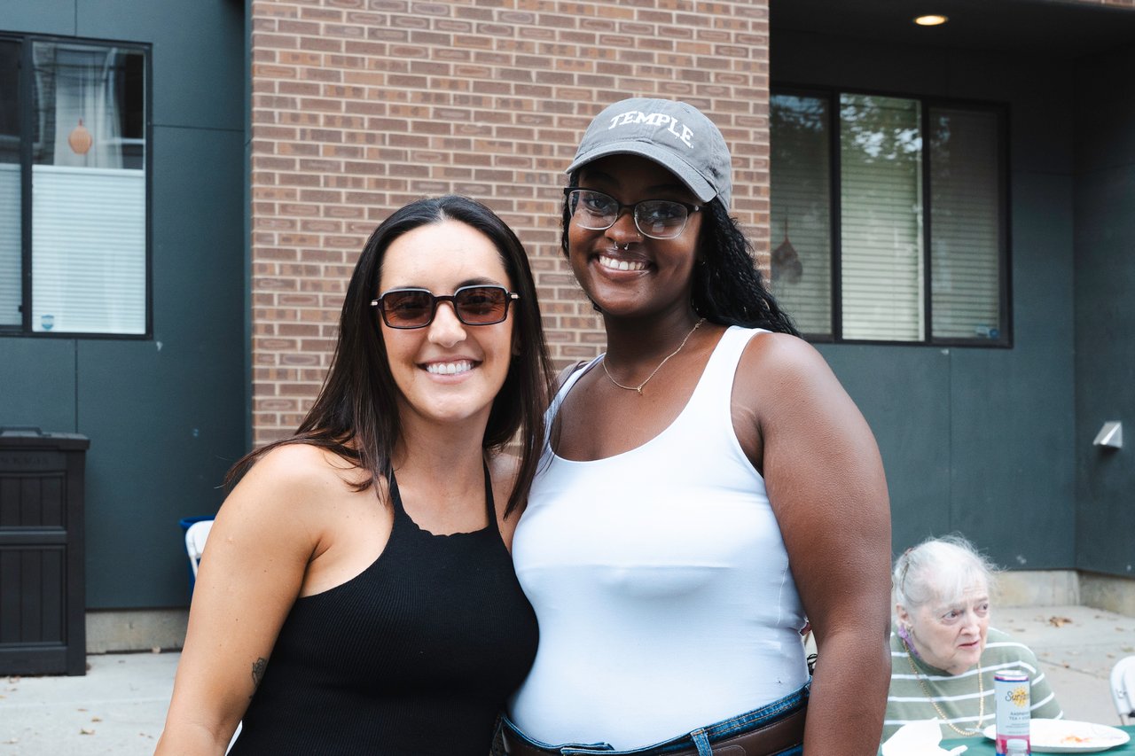 Two women pose together outside; one wears a gray Temple cap, with an older woman seated behind them.