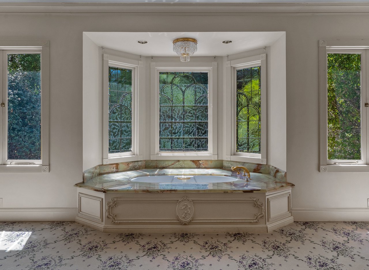 Built-in soaking tub beneath ornate stained-glass windows, with a marble surround and crystal chandelier.