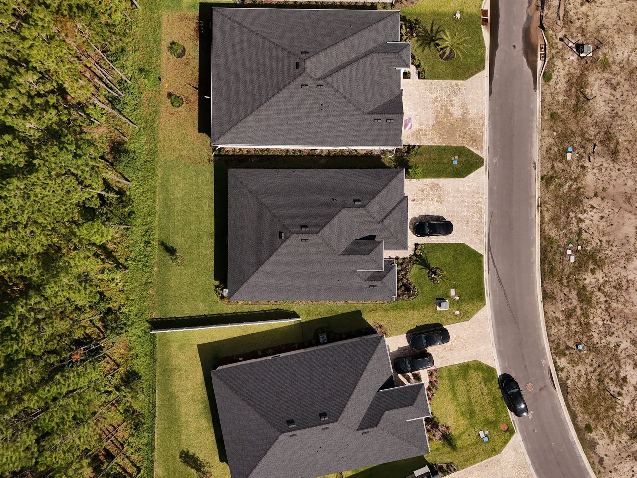 Aerial view of suburban homes with dark roofs, lawns, and driveways beside a wooded area.