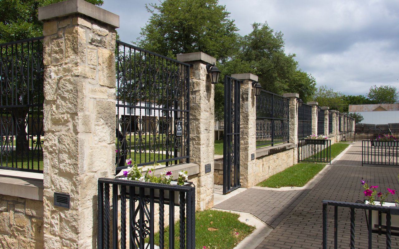 Paved walkway beside a wrought-iron fence with stone pillars, lanterns, and flower planters.