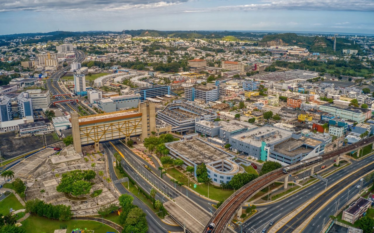 Aerial view of a city with an elevated rail line, major roads, and green hills.