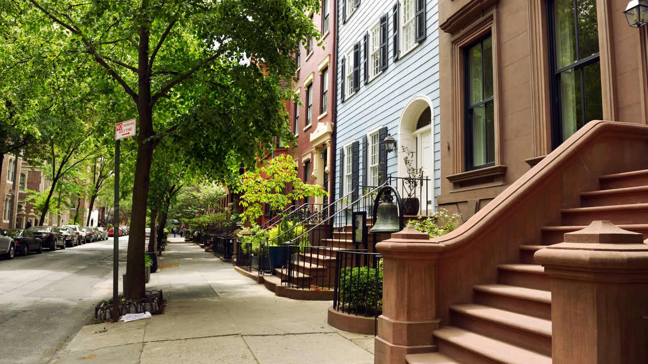 Tree-lined New York townhouse street with brownstone stoops and a pale blue clapboard facade.