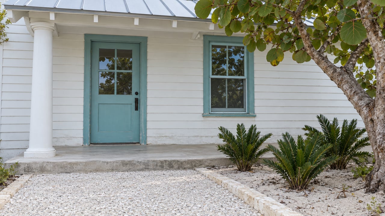 White wood cottage entrance with muted turquoise door and window frames, a shallow metal roof, and sea-grape planting beside a shell-concrete path.