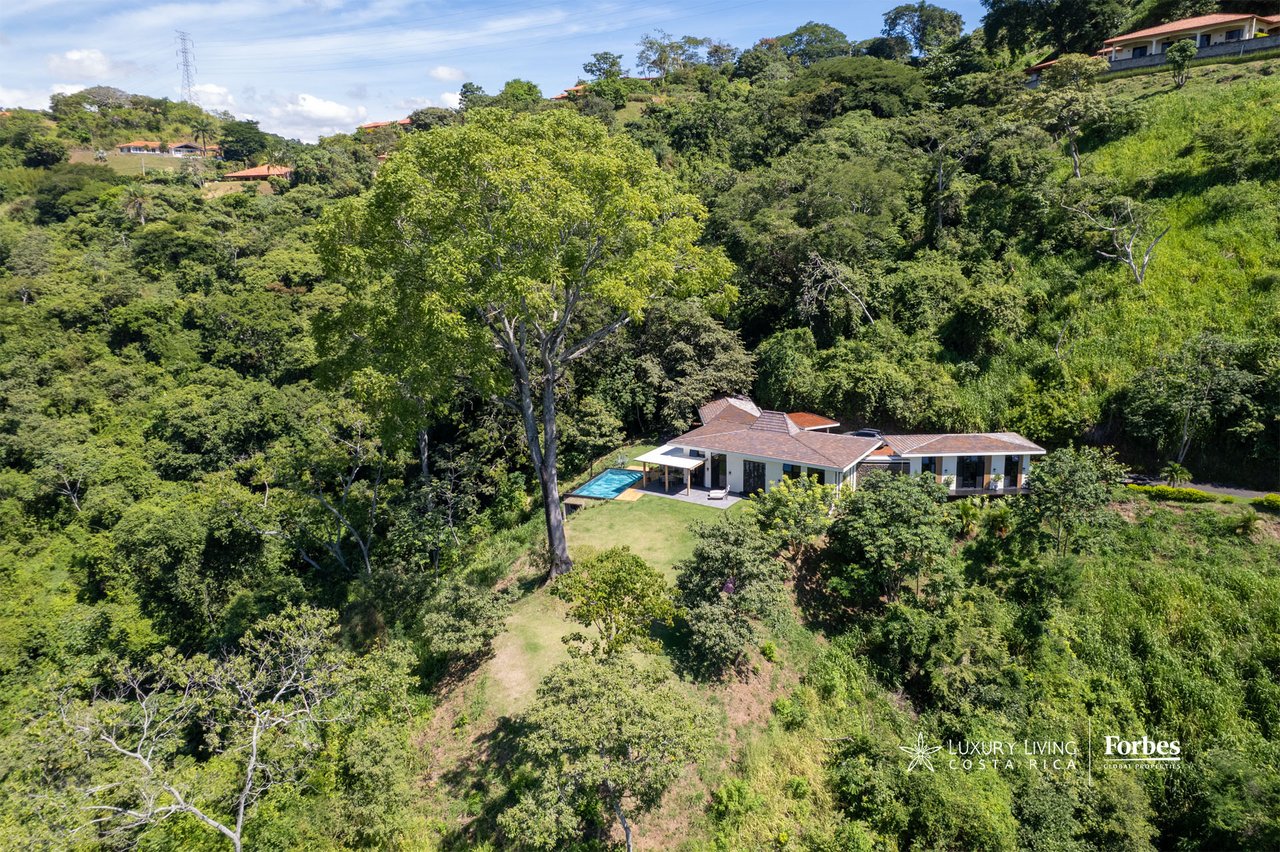 Aerial view of a modern home and turquoise pool amid lush hillside vegetation.