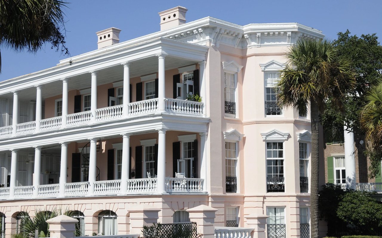 Historic pale pink building with white columns, broad balconies, and palm trees.