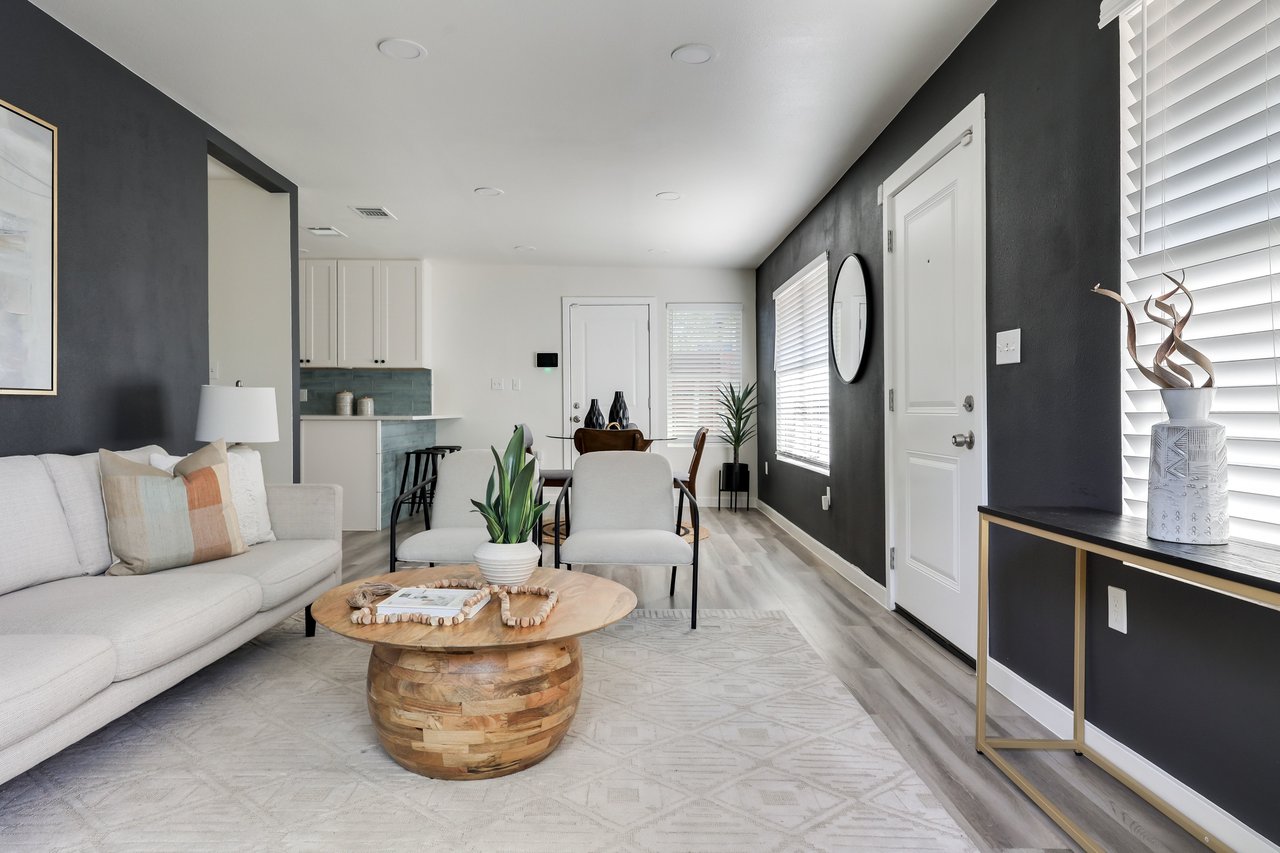 Staged living room with a white sofa, armchairs, round wood coffee table, and open kitchen.