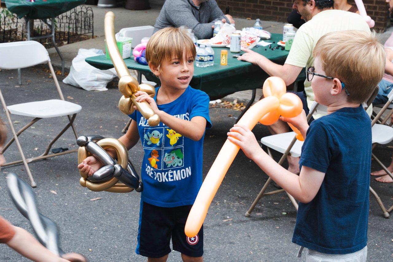 Children making balloon animals at an outdoor block party; one holds a gold-and-black balloon creation.