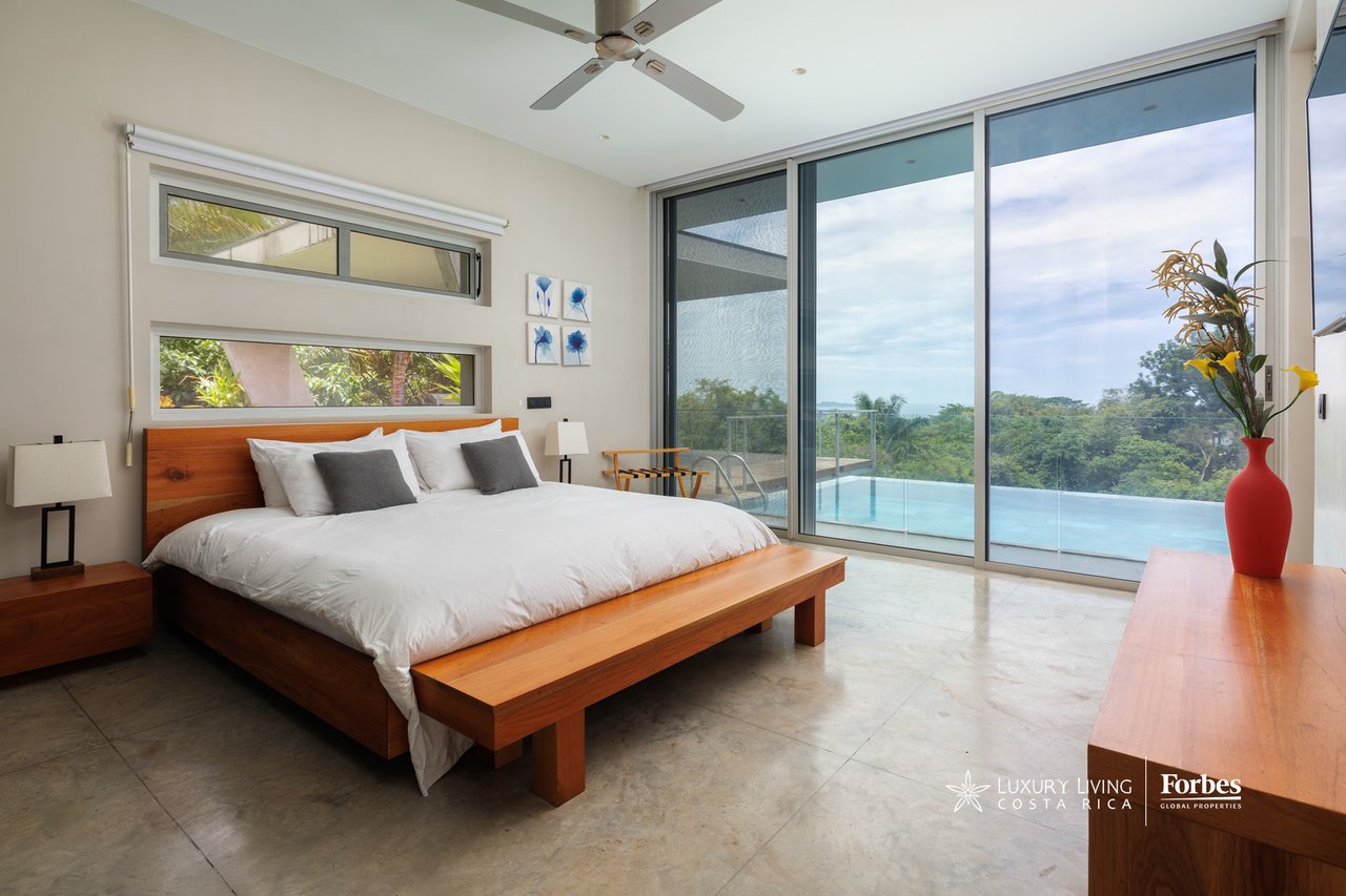 Modern bedroom with a wooden bed and glass doors overlooking a pool and tropical greenery.