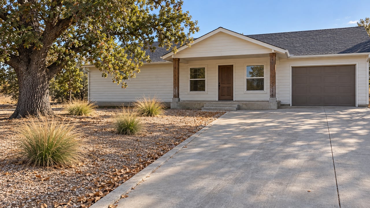 Single-story white lap-sided home with a covered timber-post porch, long concrete approach, mature valley oak and deer grass in dry gravel planting.