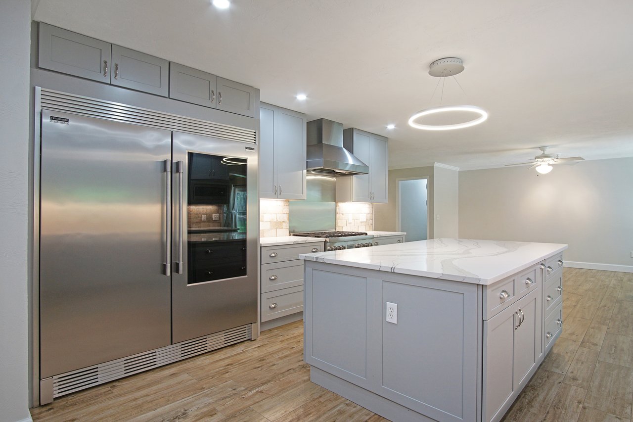 Modern kitchen with gray cabinets, large stainless steel fridge, and a spacious marble island. Circular light fixture overhead and light wood floors.
