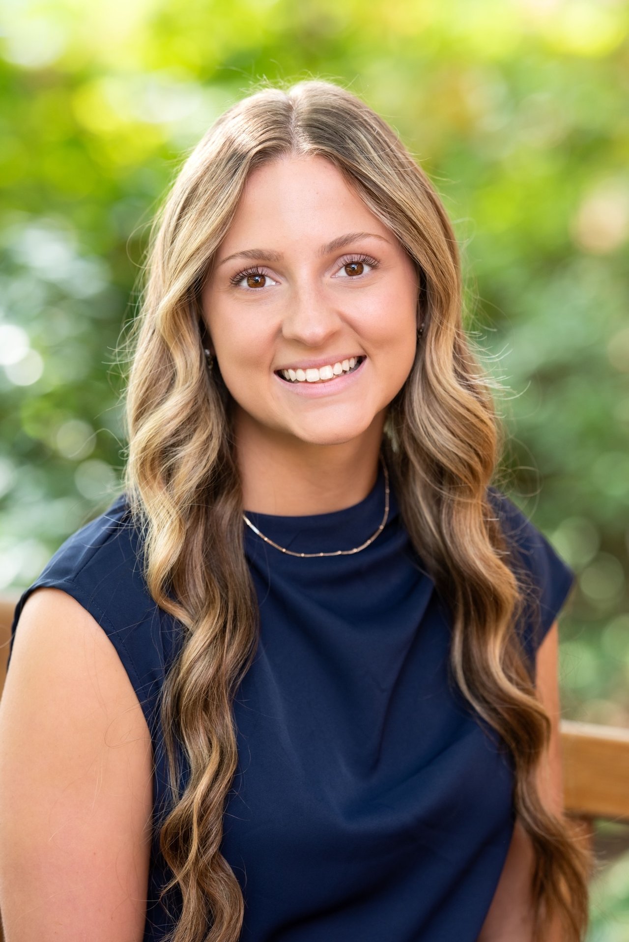 Cora Cox smiling in a navy sleeveless top against leafy greenery.