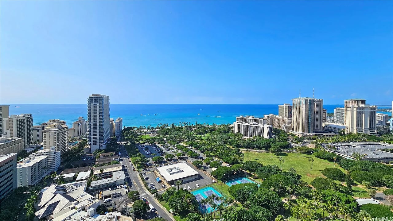 Panoramic ocean and city view from a high-rise, with green parkland and tennis courts below.