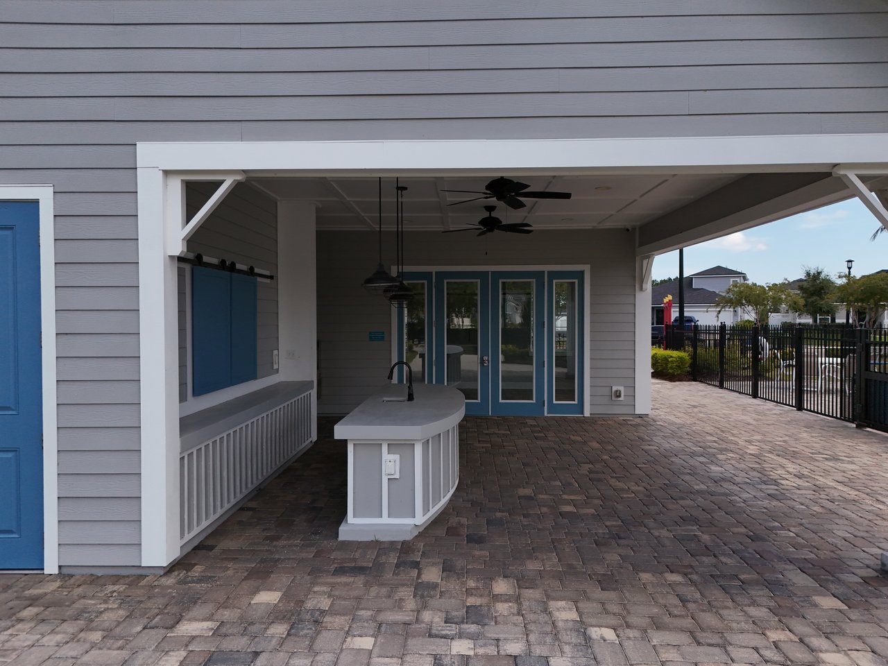 Covered outdoor pavilion with a curved sink counter, blue doors, ceiling fans, and brick paving.