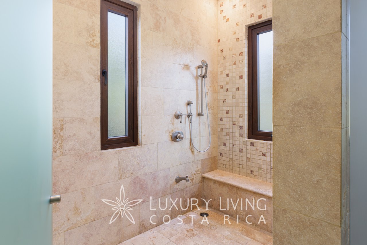 Walk-in shower with beige stone tile, mosaic accent wall, built-in bench, and narrow frosted windows.