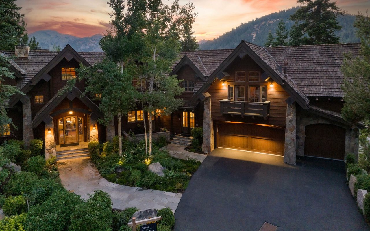 Dark-wood mountain home with stone accents and glowing windows at dusk, backed by mountains.