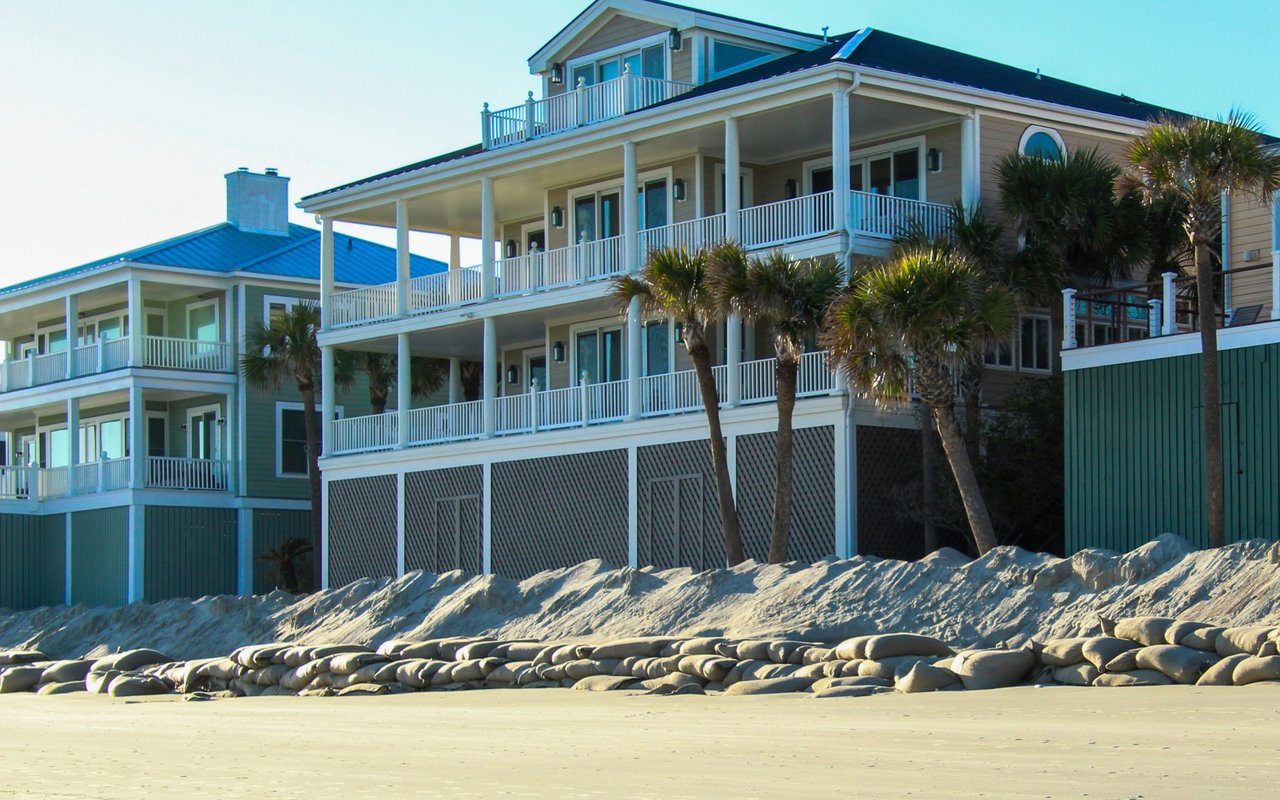 Multi-story coastal homes with white balconies and palm trees behind a rock seawall and sandy beach.