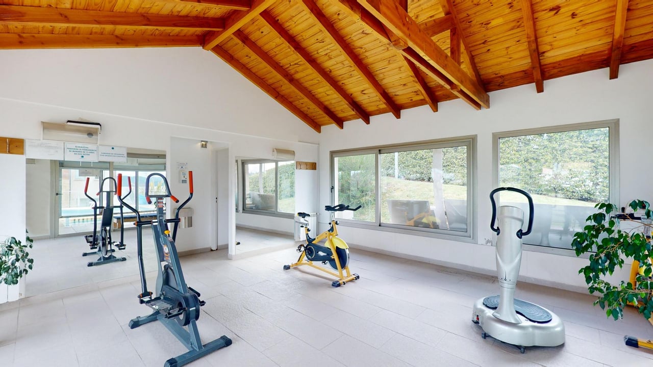 Indoor gym with exercise machines beneath a vaulted wooden ceiling and large windows.