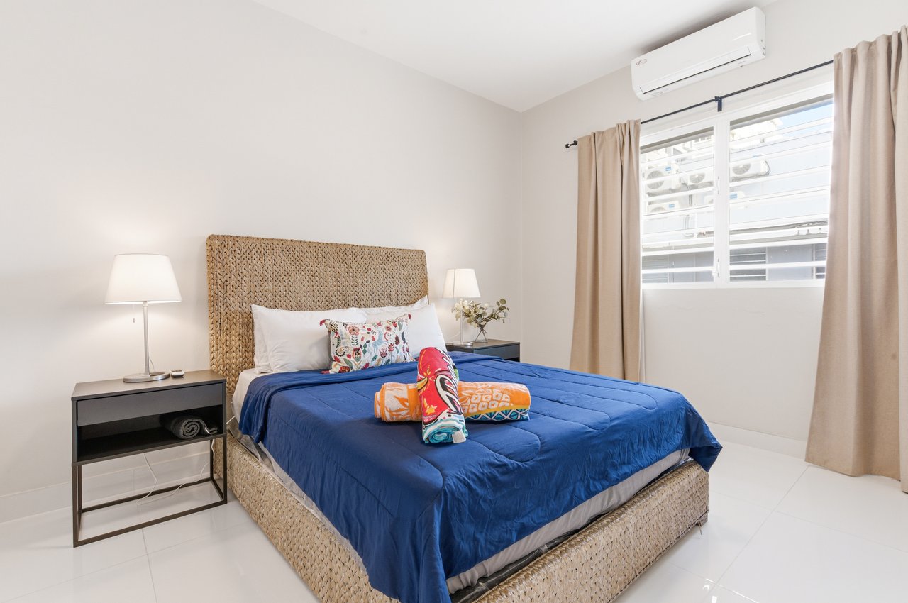 Furnished bedroom with a woven bed frame, blue bedspread, curtained window, and wall-mounted air conditioner.