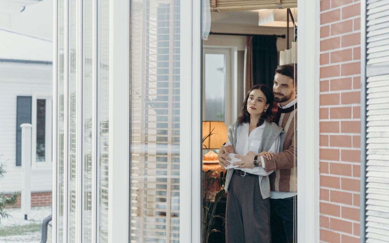 A couple embraces in their doorway, with a warmly lit room behind them and snow outside.