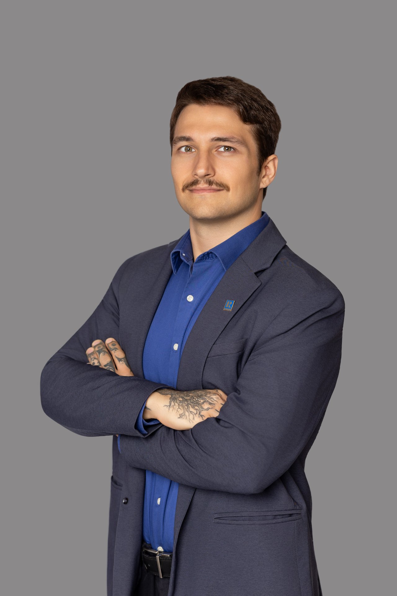 Evan Scott in a navy suit and blue shirt, standing with his tattooed arms crossed.