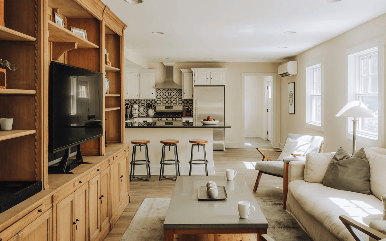 Open-plan living room and kitchen with built-in wooden shelving, a cream sofa, and a three-stool island.