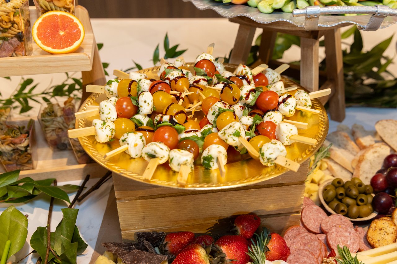 Caprese skewers on a gold platter, surrounded by fruit, olives, bread, cheese, and sliced meats.
