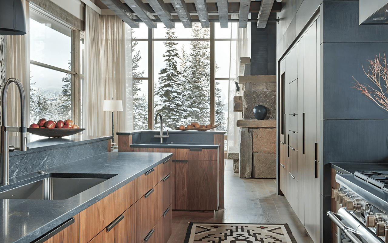 Modern kitchen with stone counters, warm wood cabinetry, and snowy trees visible through tall windows.