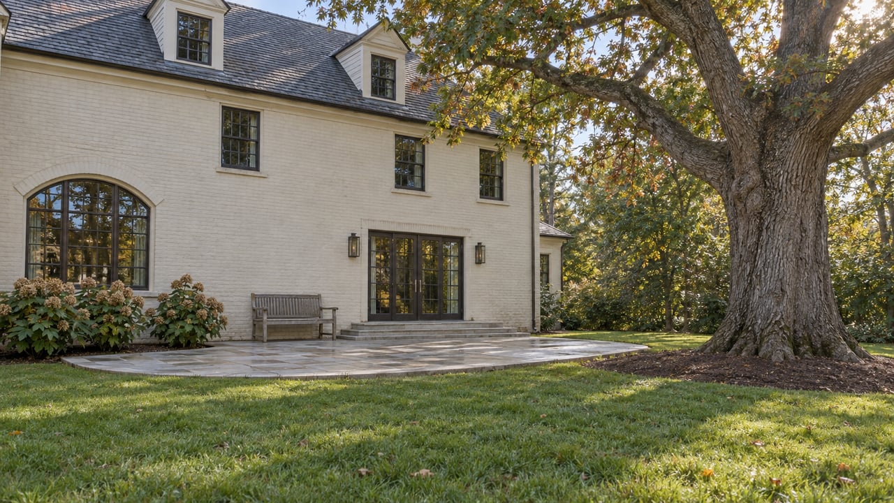 Ivory painted-brick home with a limestone rear terrace, deep lawn, and a mature white oak spreading above the garden.