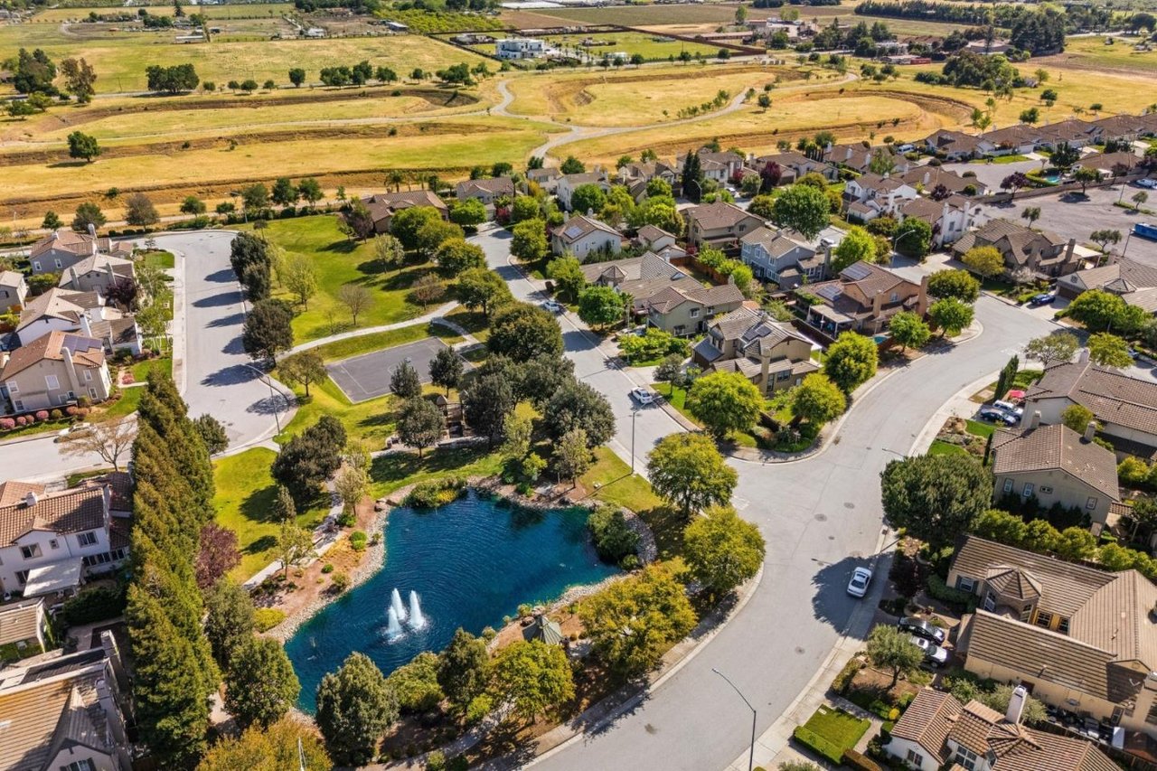 Aerial neighborhood view with homes around a fountain pond and open fields beyond.
