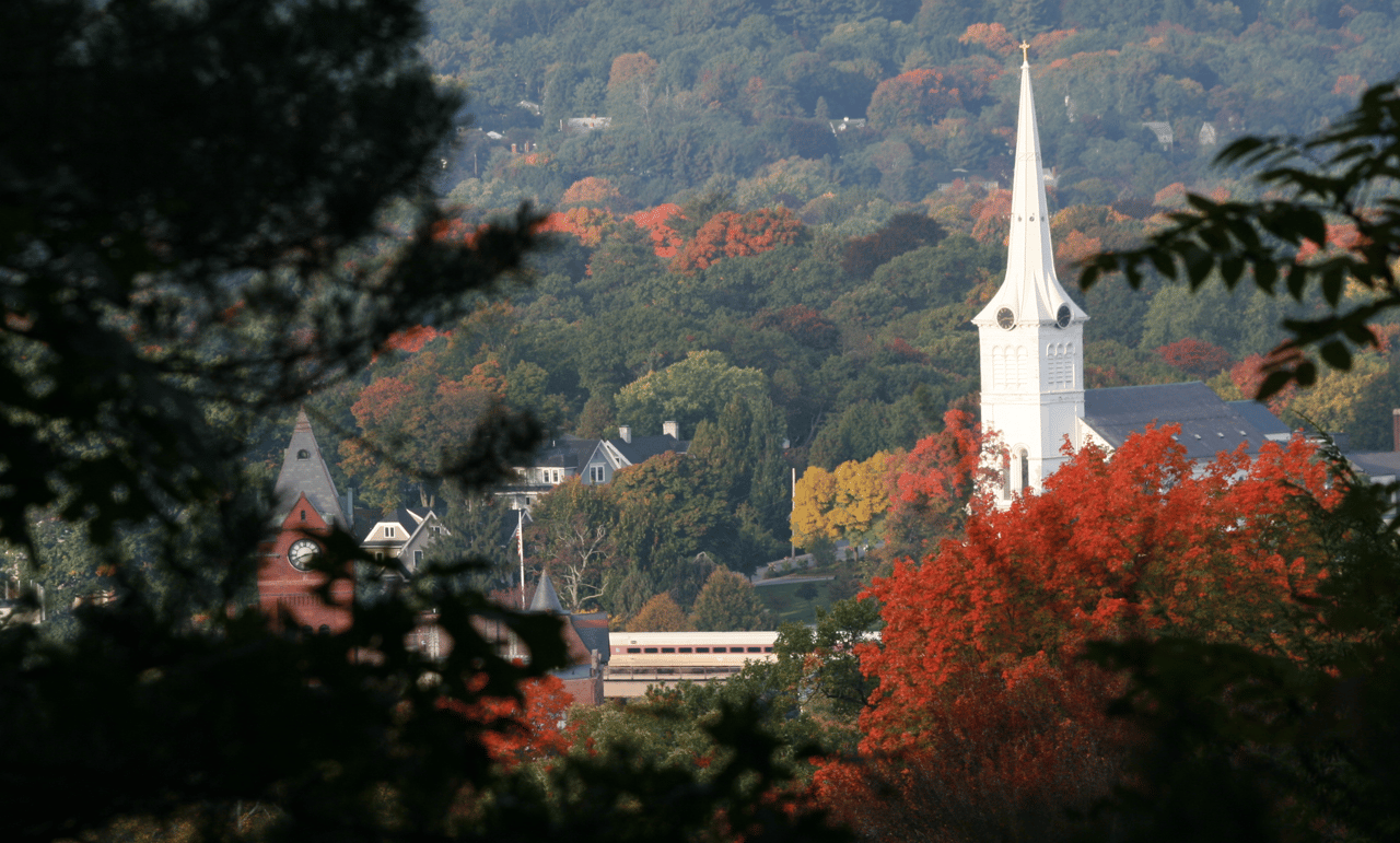A white church steeple rises above an autumn-colored town as a train passes.