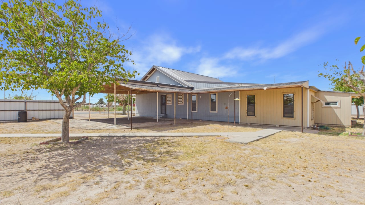 Single-story gray and tan home with a long covered carport, metal roof, and fenced yard.