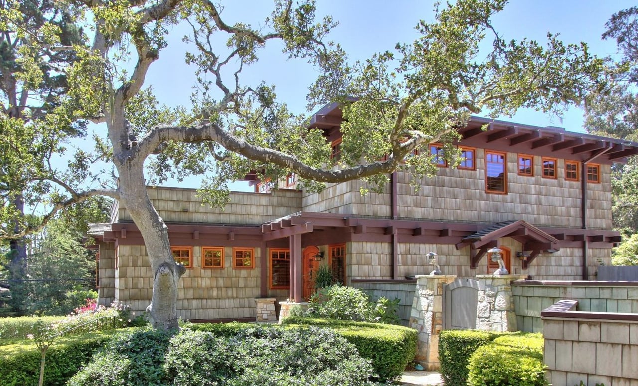 Two-story shingle-sided home with reddish trim, shaded by a large tree above clipped garden hedges.
