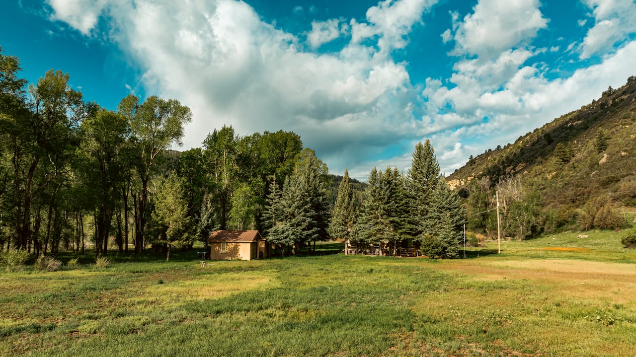 Mountain meadow with a small shed, evergreen trees, and wooded hillsides.