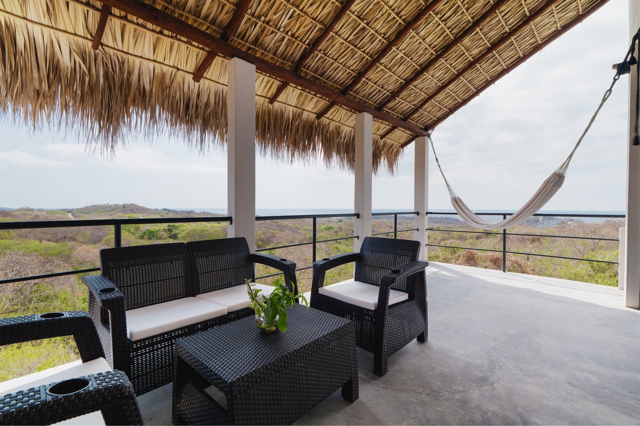 Covered terrace with wicker seating and hammock overlooking green hills.