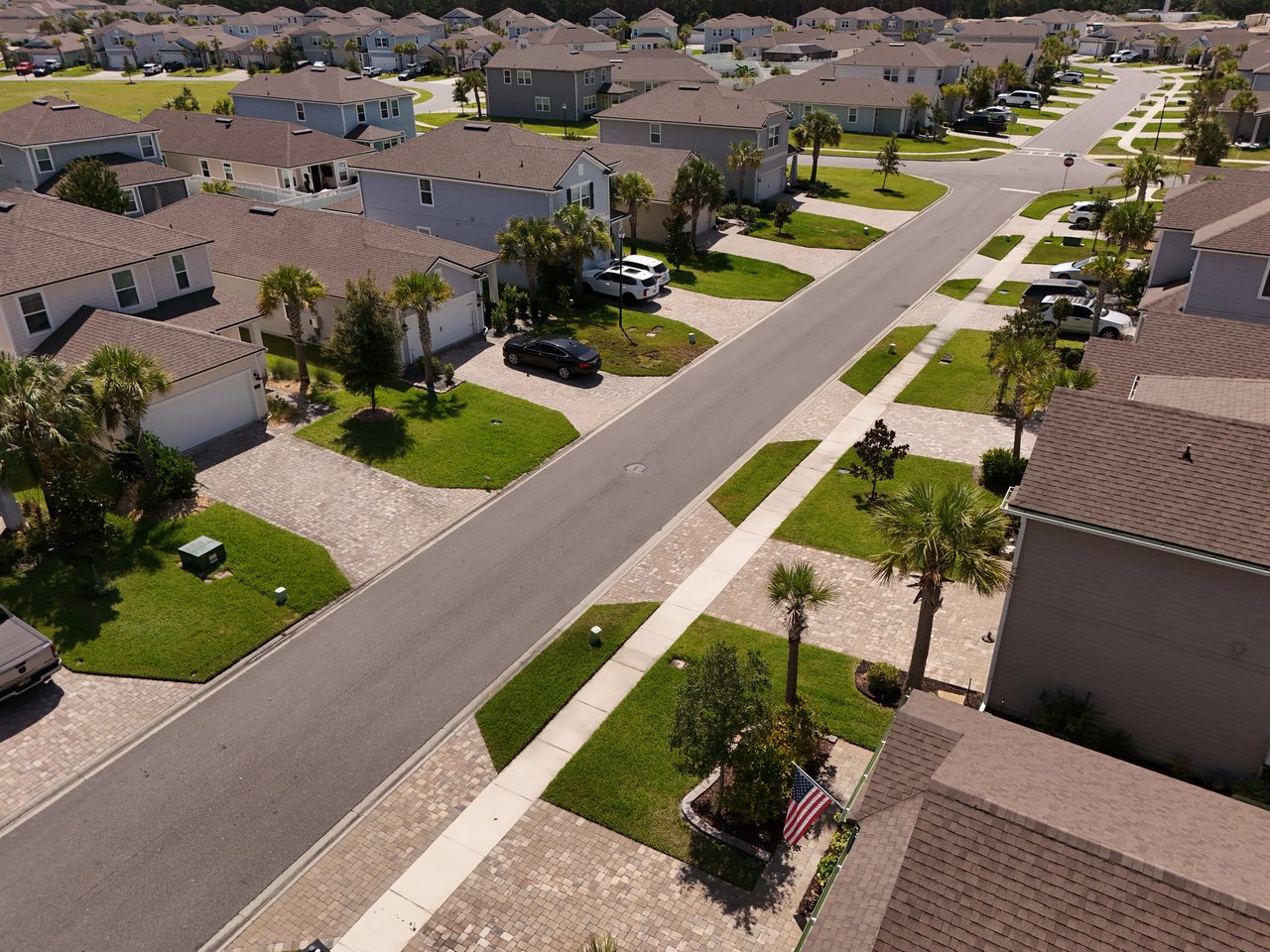 Aerial view of homes along a neighborhood street, with palm trees, green lawns, and an American flag.