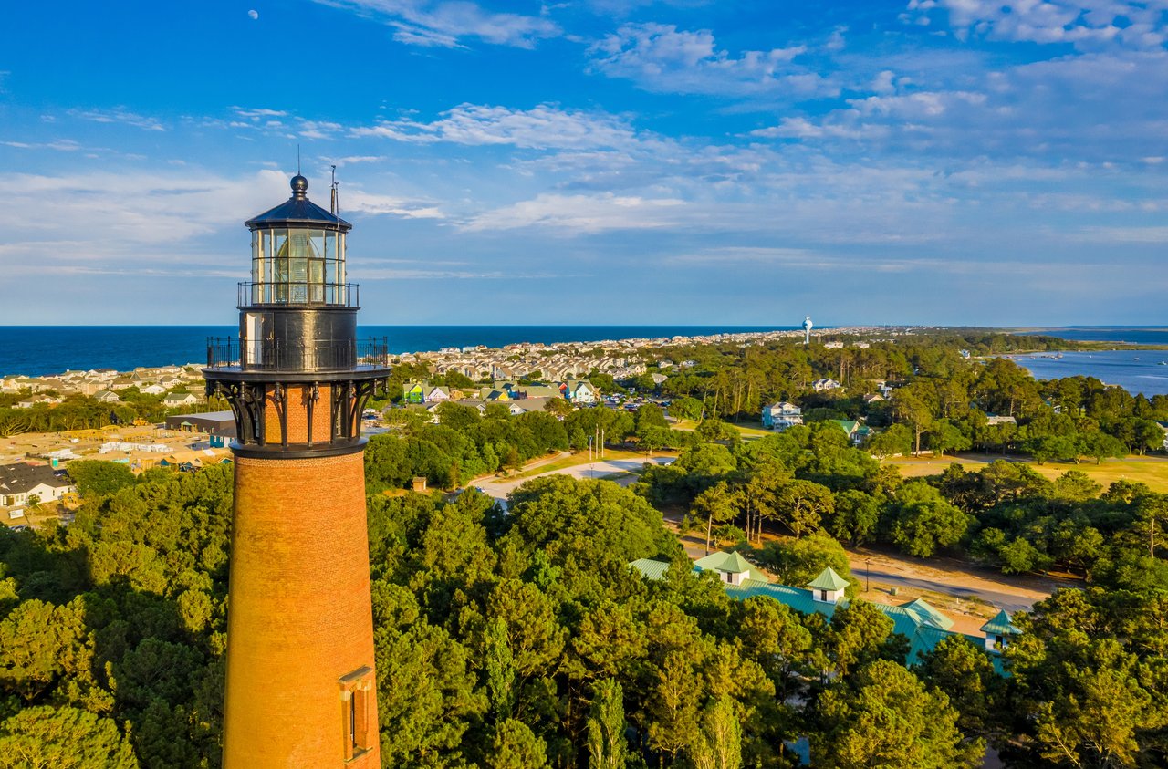 Aerial view of a brick lighthouse above coastal homes, trees, and ocean.